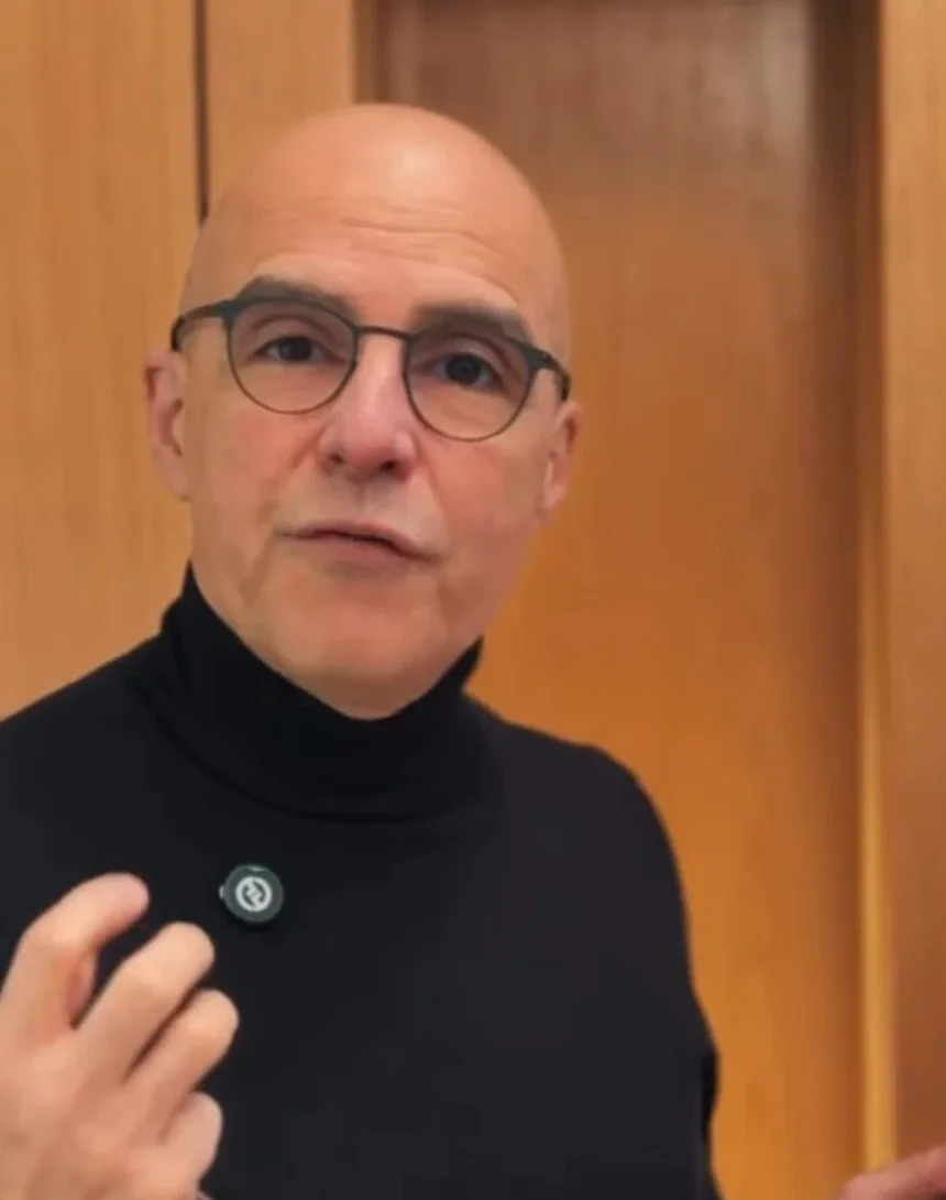 Dr. Afrânio Bernardes, de gola alta preta e óculos, explicando com a mão em gesto diante da parede de madeira do consultório