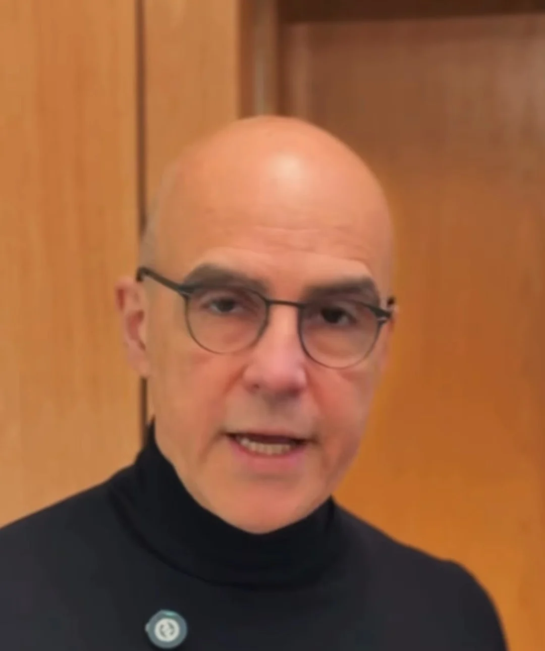 Dr. Afrânio Bernardes, de gola alta preta, falando para a câmera diante de uma parede de madeira
